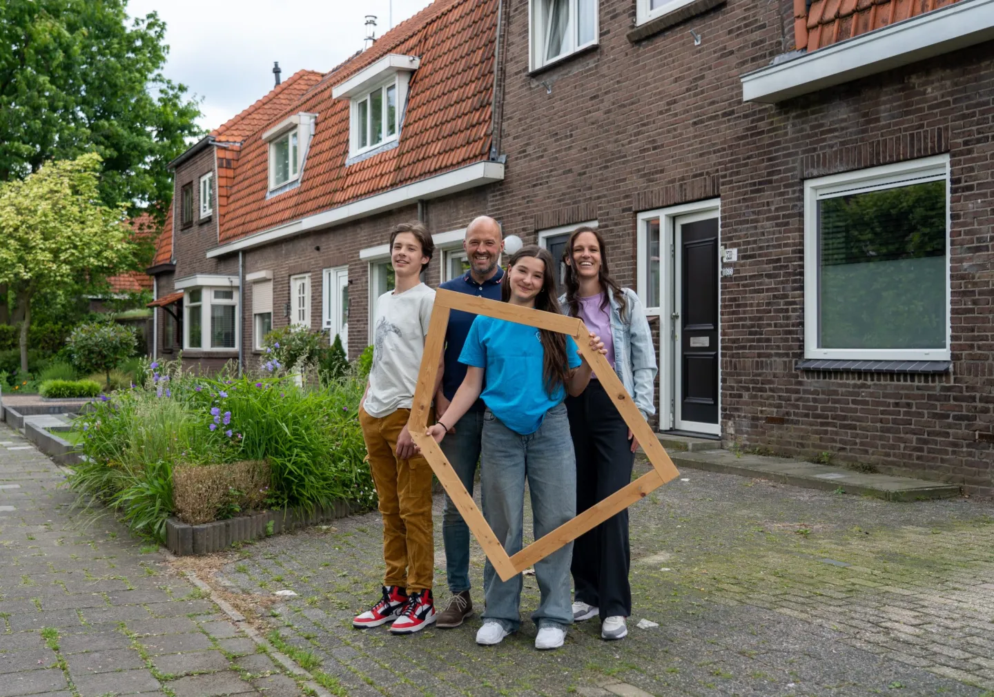 Een vrouw, een man, een jongen en meisje staan voor een rij met huizen. Meisje heeft een groot houten frame van een huis in haar handen. 