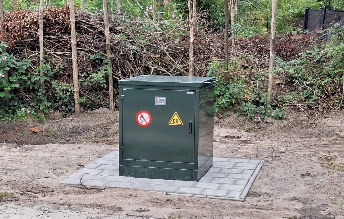 Groen transformatorhuisje omringd door zand, met op de achtergrond struiken