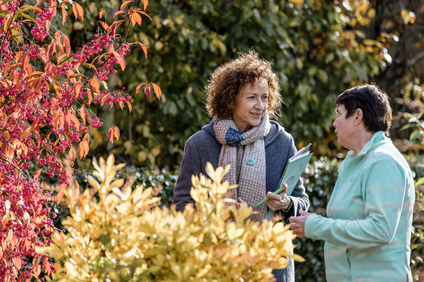 Twee dames in gesprek in een tuin, met daarbij een geel en rood struikje