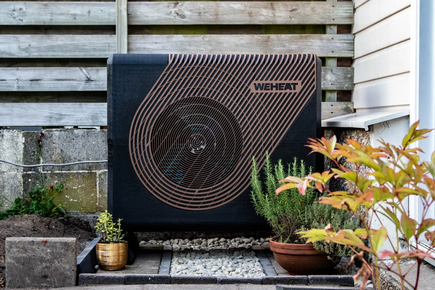 Zwarte buitenunit van warmtepomp tegen een houten schutting
